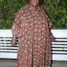 A woman outside standing against greenery wearing a raisin floral tapestry maxi caftan with three quarter balloon sleeves .