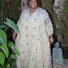A woman wearing a maxi caftan with a floral pattern in a color that could be described as deep beige ground with tan, green and black paisley tapestry, standing outdoors.