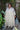 Woman wearing a beige ground turquoise flower tapestry caftan standing in a garden with plants and a statue.