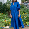 A woman standing in a garden wearing a blue maxi dress with balloon sleeves and wide elastic cuffs, made of silk, posing next to a building with a blue sign on it.