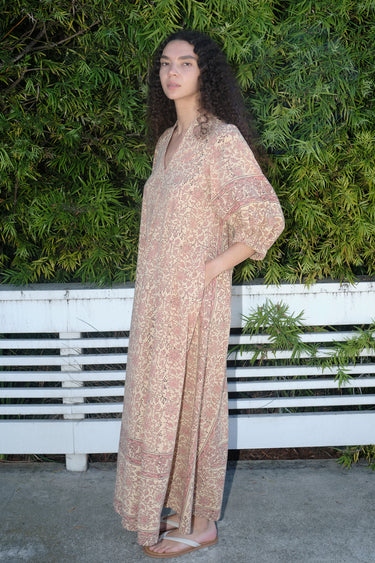 A woman standing outdoors wearing a blush maxi dress with floral hand block print design, featuring balloon sleeves and wide elastic at the cuffs, side view.