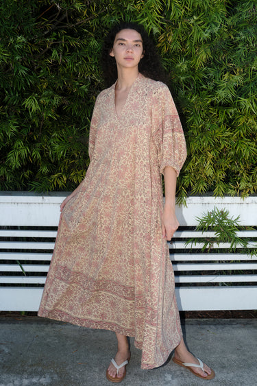 A woman standing outdoors wearing a blush maxi dress with floral hand block print design, featuring balloon sleeves and wide elastic at the cuffs.