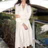 A woman standing outdoors by a canal wearing a winter white dress cut like a caftan with long full sleeves. 