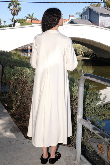 A woman standing outdoors by a canal wearing a winter white dress cut like a caftan with long full sleeves, back view.