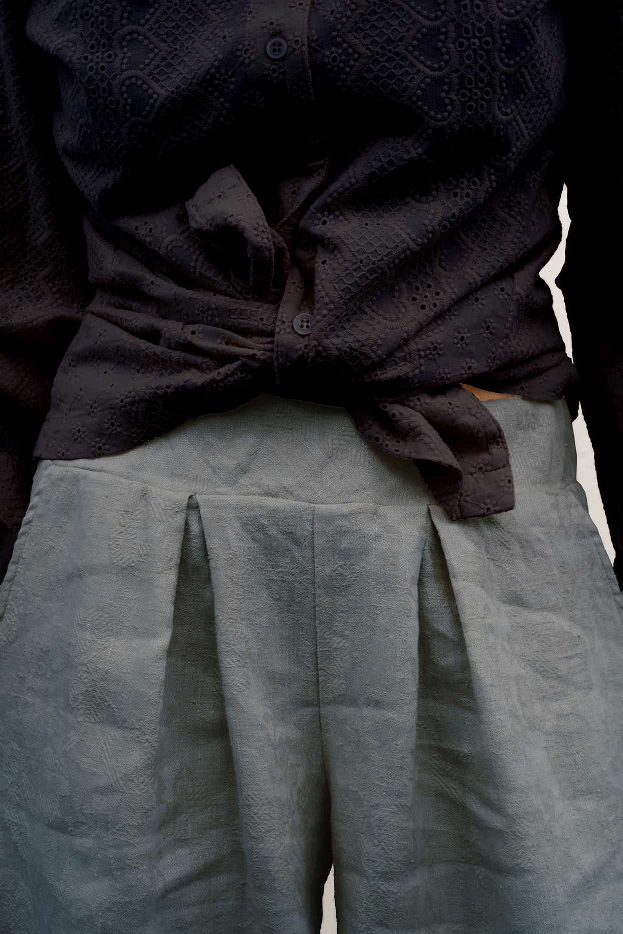 Close-up of a person wearing a dark charcoal textured button-up shirt tied at the waist and light gray pleated trousers.