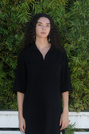A woman standing in front of greenery wearing an a black maxi shirtdress with a collar, sleeves, and a side slit, with a body made of crinkle textured osnaburg cotton.