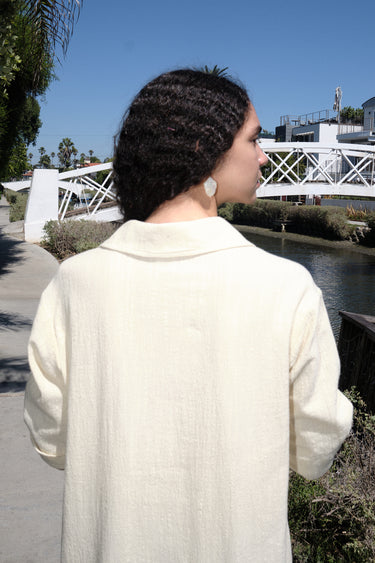 A woman standing on a pathway wearing an alabaster beige maxi shirtdress with a collar, sleeves, and a side slit, with a body made of crinkle textured osnaburg cotton, back view of collar. 