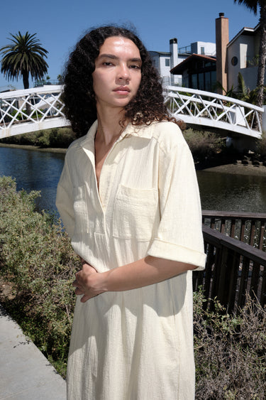 A woman standing on a pathway wearing an alabaster beige maxi shirtdress with a collar, sleeves, and a side slit, with a body made of crinkle textured osnaburg cotton. Showing collar and placlet details. 