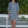 A person standing by a white fence with greenery wearing a mini shirt dress with a shirt collar and an "xo" Ikat pattern.