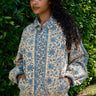 Woman wearing a blue and beige floral patterned outfit  standing against a green leafy background