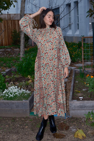 Woman standing in garden wearing a long sleeve midi length dress in an ecru with red flower tapestry.