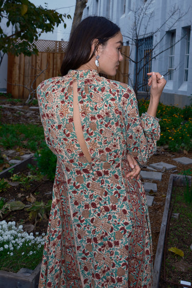 Woman standing in garden wearing a long sleeve midi length dress in an ecru with red flower tapestry, back view.
