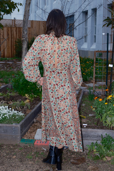 Woman standing in garden wearing a long sleeve midi length dress in an ecru with red flower tapestry, back view.