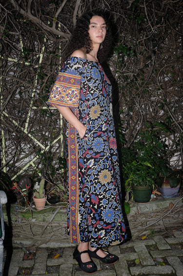 Woman standing in garden wearing a maxi dress with a bardot neckline, cut in a black ground sunflower tapestry. 