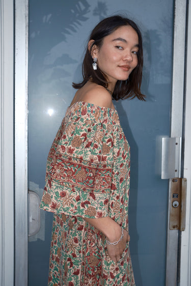 A woman standing in front of a door, wearing an off-the-shoulder floral tapestry dress with pockets.