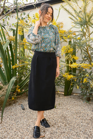 Woman in a floral blouse and black skirt standing in a garden with yellow flowers and cacti.
