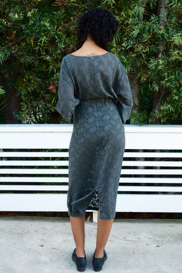 Person wearing a gray lace outfit sitting on a white bench with greenery in the background