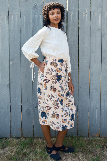 Woman wearing a white blouse and floral skirt standing against a wooden fence.