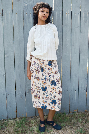 Woman wearing a white blouse and floral skirt standing against a wooden fence.