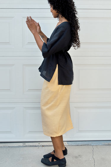 Person wearing ablack tee and yellow skirt against a white wall.