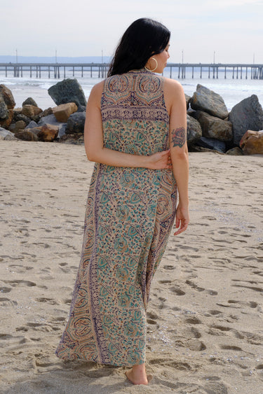 A woman at the beach wearing a sleeveless maxi caftan in a beige ground tapestry, turquoise flower handloom, back view.