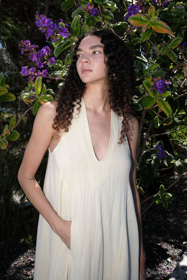 A woman outside wearing an alabaster off white maxi caftan with deep cut arm holes and bias cut sides, featuring front cell phone pockets.