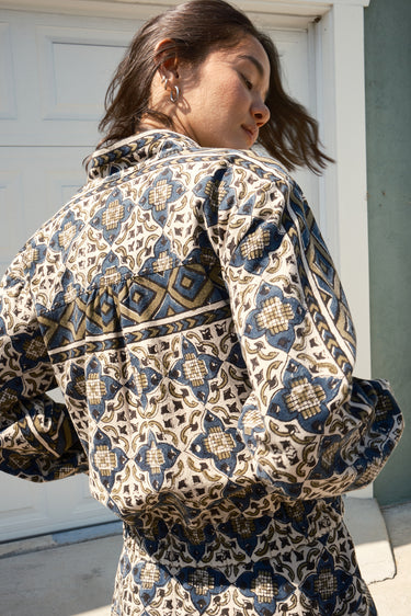 A woman standing against garage wearing a cropped, patterned tapestry jacket with a zip front and elastic sleeves. back view