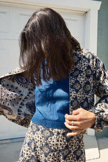 A woman standing against a garage wearing a cropped, patterned tapestry jacket with a zip front and elastic sleeves. She is s showing the lining.