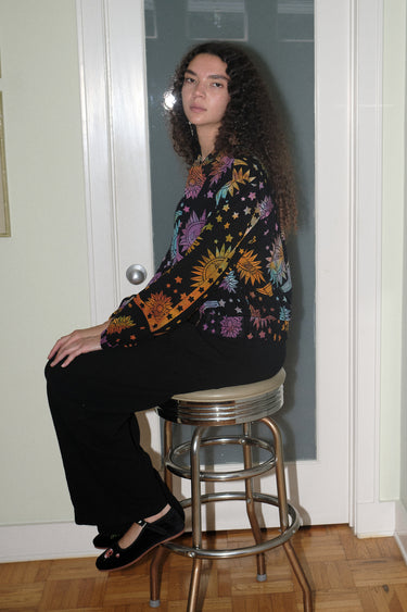 A woman wearing a cropped black ground with rainbow suns, moons and stars tapestry jacket with a zip front, standing in a home environment.