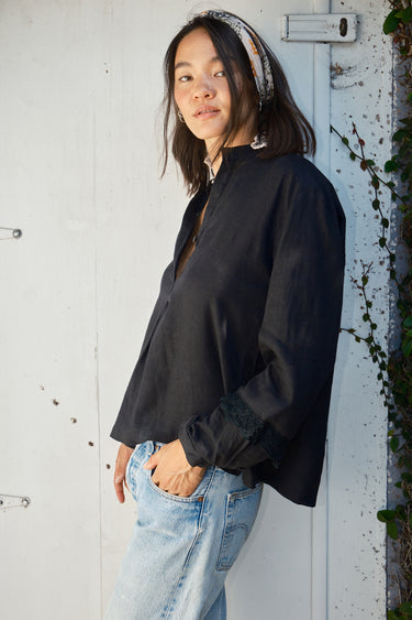 A woman wearing a black linen blouse with long sleeves and a band collar, paired with denim jeans.