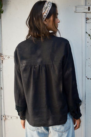 A woman wearing a black linen blouse with long sleeves and a band collar, paired with denim jeans, back view