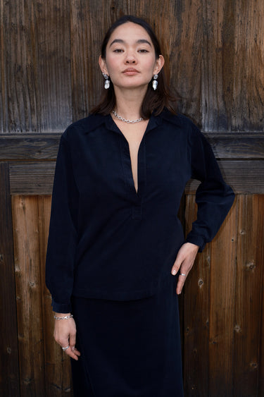 A woman standing by a brown wood fence wearing an ebony black  corduroy shirt with long sleeves.