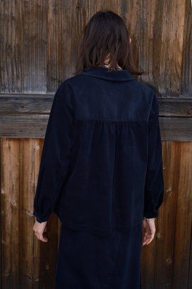 A woman  standing by a brown wood fence wearing an ebony black  corduroy shirt with long sleeves, back view. 
