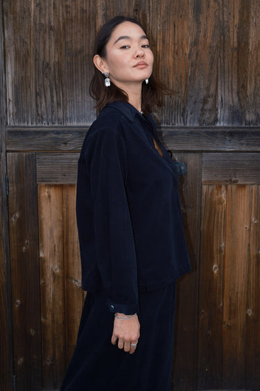 A woman standing against a wood fence wearing an ebony black corduroy shirt with long sleeves.