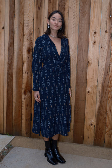 A woman standing in front of a wooden gate wearing black ikat shirt dress with long sleeves and an obi belt.