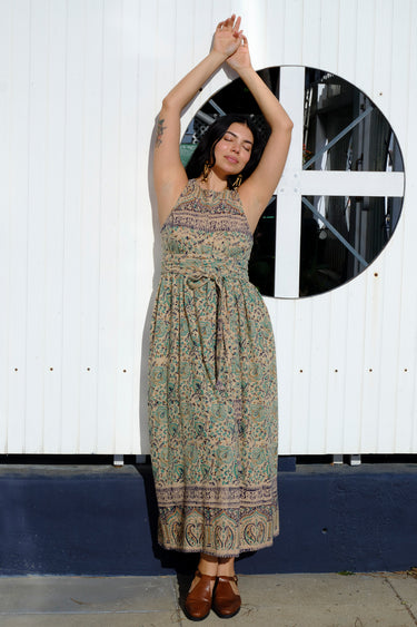 A woman standing by a white fence wearing a beige ground turquoise flower tapestry wrap maxi dress.