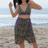 
A woman standing at the beach wearing a floral tapestry  wrap skirt cut using both green and blue tapestry of the same print tapestry mixed together. Worn with a matching bra top.