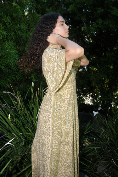 woman standing in garden wearing a light green print tapestry dress with zip front, v neck and elbow length sleeves, side view.