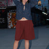 A woman standing on the sidewalk wearing a raisin colored Bermuda short, long sleeve top and a pair of strappy sandals. The background includes graffiti and a building.
