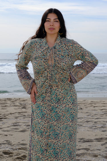 A woman standing at the beach wearing tan with green turquoise flower tapestry shirt with bell sleeves and a matching skirt. 
