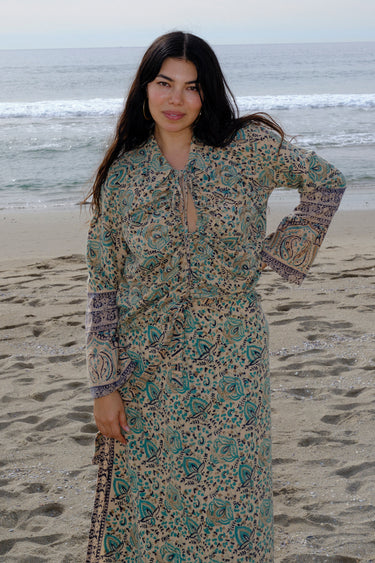 A woman standing at the beach wearing tan with green turquoise flower tapestry shirt with bell sleeves and a matching skirt. 