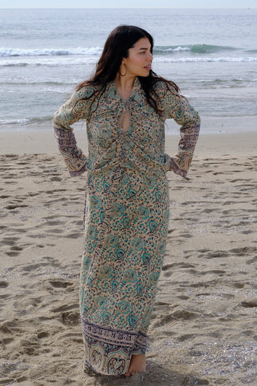A woman standing at the beach wearing tan with green turquoise flower tapestry shirt with bell sleeves and a matching skirt. 