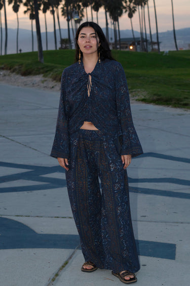 A woman standing at Venice Beach at dusk wearing dark blue midnight sky handloom tapestry shirt with bell sleeves and matching trousers.