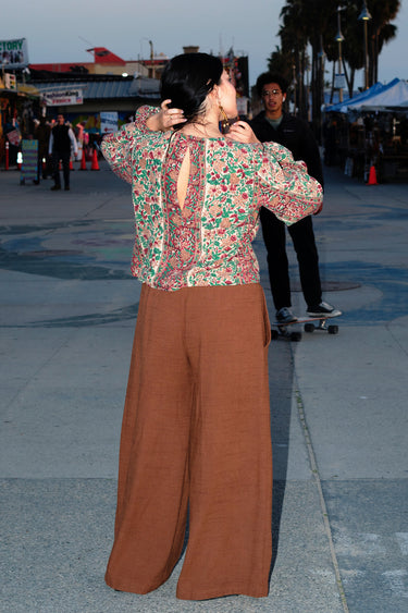 A woman standing at a beach boardwalk wearing a nutmeg brown trouser and a floral print blouse. Back View