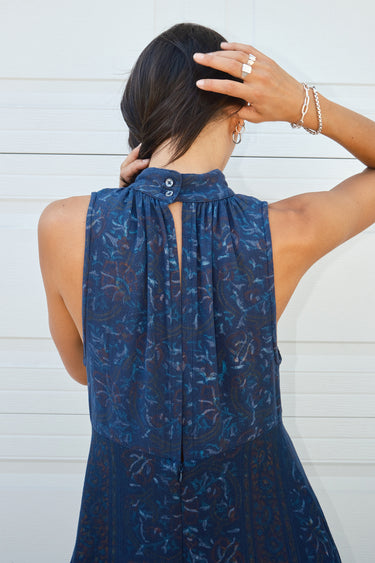 Back View of a woman standing in front of a white garage door, wearing a navy dress with a band collar, empire waist, and hand woven, hand block printed Indian Tapestry fabric. 