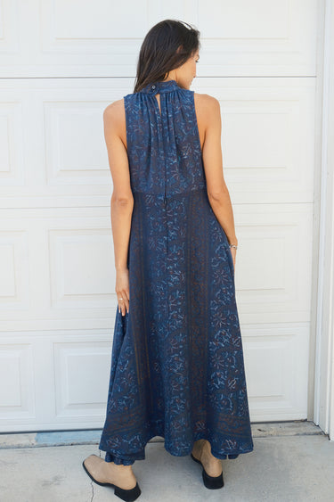 A woman standing in front of a white garage door, wearing a navy dress with a band collar, empire waist, and hand woven, hand block printed Indian Tapestry fabric. She is wearing tan boots. 
Back view 