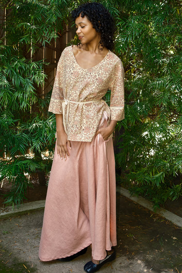Woman wearing a gold top and pink skirt standing in front of green foliage.