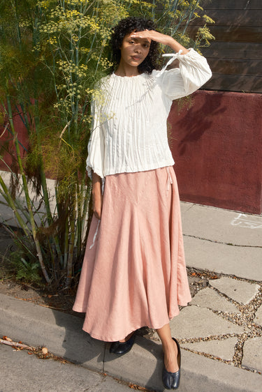 Woman wearing a white blouse and pink skirt standing outdoors.