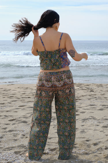 A woman standing at the beach wearing a floral tapestry camisole cut using both green and blue tapestry of the same print tapestry mixed together.Worn with matching pants. Back View