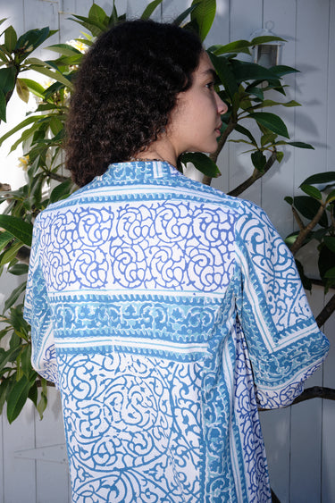 A woman standing on a pathway with a white and blue floral print maxi dress with three-quarter sleeves, back view.